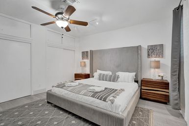 Modern neutral master bedroom with an upholstered king bed and tall ribbed headboard, wooden nightstands with lamps, ceiling fan, patterned area rug, and folded towels on the bed — cozy, minimalist interior.