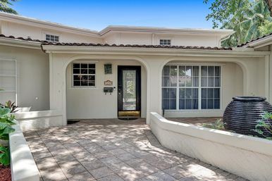 Sunny Mediterranean-style stucco home entrance with clay tile roof, arched covered porch, black glass front door, large grid window, paver courtyard and curved planter featuring a round dark water urn and tropical palms.