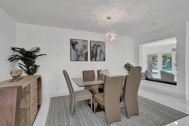 Bright modern dining room with glass-top table and six taupe chairs, a starburst pendant chandelier, abstract wall art, wood sideboard and potted plants, open passthrough showing poolside seating and windows.