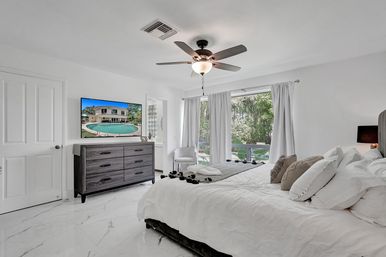 Bright modern white master bedroom with king bed, marble-look floors, ceiling fan, dark wood dresser and wall-mounted TV, large windows with white curtains overlooking lush tropical backyard foliage.
