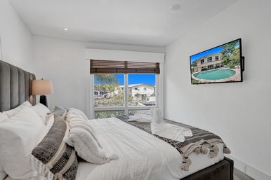 Bright modern bedroom with a plush bed layered in textured pillows and a cozy throw, large window framing a palm-tree and neighboring homes, wall-mounted TV, crisp white walls and natural light.