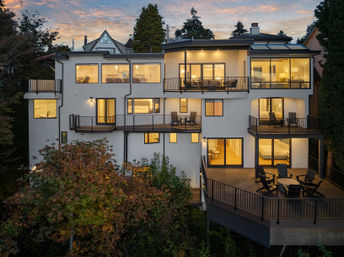 Sunset view of a modern multi-level hillside home with warm-lit interiors, large windows, multiple balconies and a spacious deck with Adirondack chairs overlooking treetops.