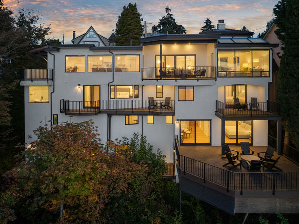 Sunset view of a modern multi-level hillside home with warm-lit interiors, large windows, multiple balconies and a spacious deck with Adirondack chairs overlooking treetops.