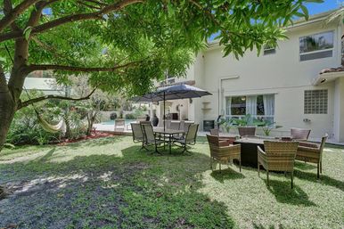 Shaded suburban backyard patio with umbrella-covered dining table, wicker seating around a fire pit, hammock under a tree, green lawn and pool beside a two-story house