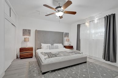 Bright modern master bedroom with gray upholstered bed, patterned rug, wooden nightstands and lamps, ceiling fan, and large curtained window — clean, minimalist interior design