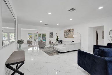 Sunlit modern open living room with glossy white marble floors, white sectional sofa, navy armchairs, grey area rug, abstract wall art, and glass doors opening to a backyard pool.