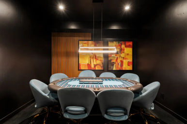 Modern private poker room interior with oval poker table and blue velvet swivel chairs, wood‑slat accent wall, warm abstract paintings and linear pendant lighting.