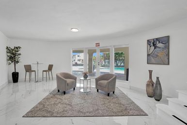 Bright modern lounge with two cozy gray armchairs on a textured rug, glass side table and white marble floors, large patio doors overlooking an outdoor pool, bar table with stools, potted plant, vases and abstract wall art.