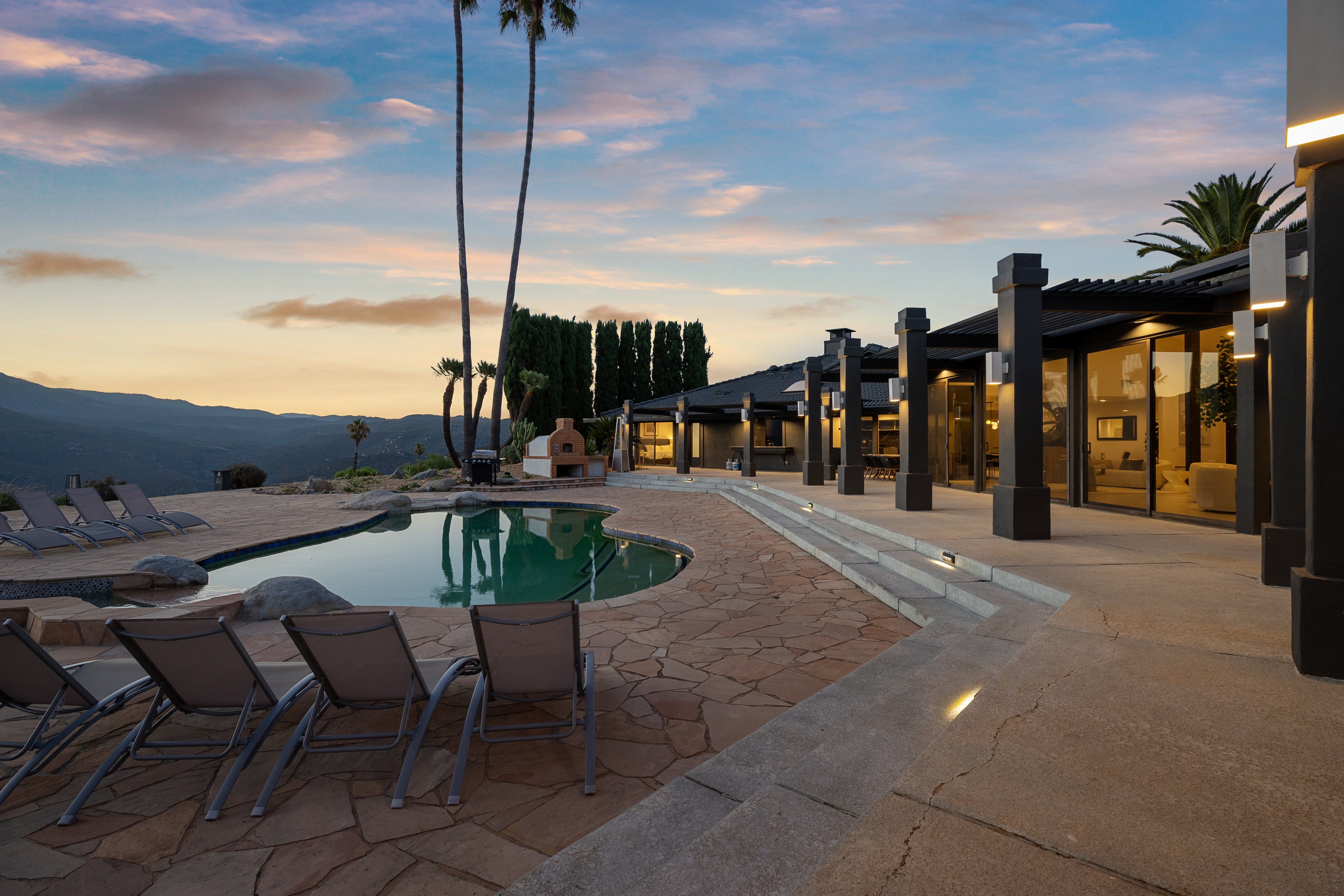 Modern luxury hillside home patio at sunset with a curved swimming pool, lounge chairs, outdoor fireplace and mountain views