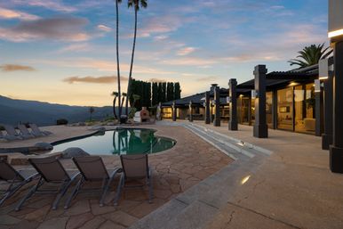 Modern luxury hillside home patio at sunset with a curved swimming pool, lounge chairs, outdoor fireplace and mountain views