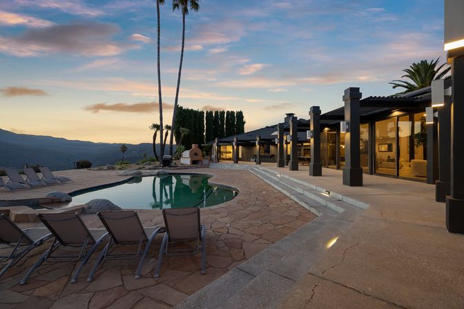 Modern luxury hillside home patio at sunset with a curved swimming pool, lounge chairs, outdoor fireplace and mountain views