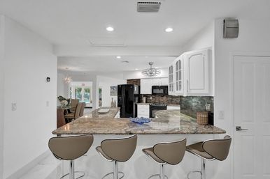Open-concept modern kitchen with granite peninsula, four tan bar stools, white cabinets, black appliances, mosaic tile backsplash, and backyard pool view.