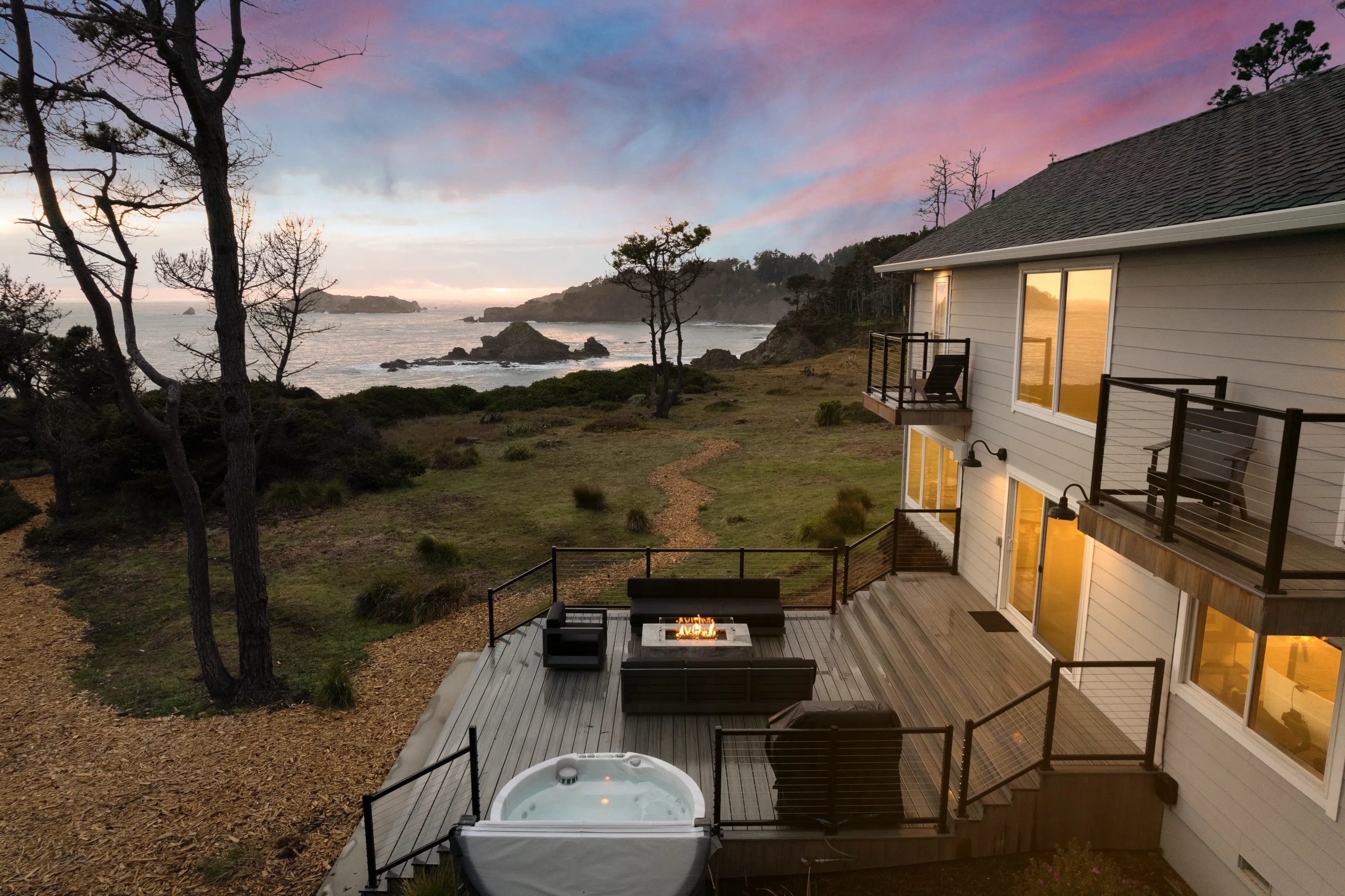 Cozy oceanfront home at sunset with a wooden deck, bubbling hot tub, lit fire pit and patio seating, balconies overlooking a winding path to a rocky coastline beneath a pink-and-blue sky.