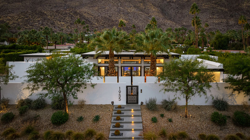 Dusk-lit modern desert home with white stucco walls and gated courtyard, illuminated stepped pathway with barrel cacti, palm trees framing a glass-front living area, xeriscape gravel landscaping and rugged mountain backdrop.