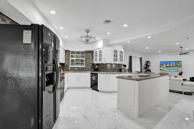 Bright open-concept modern kitchen in a family home with white cabinetry, large island with granite top, stainless refrigerator and appliances, marble-look tile floor, gray stone backsplash, recessed lighting and view into the living area.