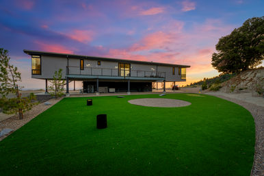 Modern elevated home on stilts overlooking a synthetic-turf backyard putting green with a gravel center, set against a pink and purple sunset sky on a hillside.