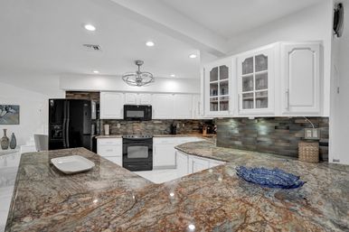 Bright modern kitchen with L-shaped polished granite countertops, white cabinetry with glass-front uppers, black appliances, gray mosaic tile backsplash, recessed lights and a stylish pendant over the breakfast bar