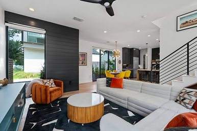 Bright modern open-concept living room and kitchen with white sectional, orange velvet chair, round wood-and-marble coffee table, black accent wall, yellow dining chairs, and sliding glass doors.