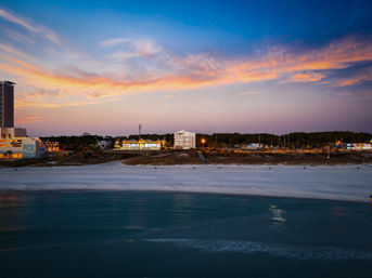Pastel sunset over turquoise ocean and wide white-sand beach, lit beachfront buildings and tree-lined coast at dusk.