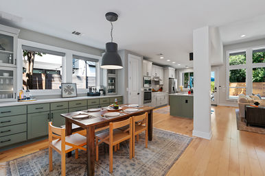 Bright modern open-plan kitchen and dining area in a suburban home with sage-green cabinets and island, wooden farmhouse table set for four, large windows overlooking a sunny fenced backyard, hardwood floors, pendant lighting and adjacent living room.