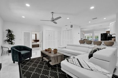 Bright modern open-plan living room with white sectional sofa, black patterned rug, wooden coffee table, green velvet armchair, kitchen bar stools and ceiling fan