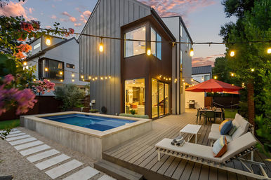 Cozy modern two-story home backyard at sunset with a raised plunge pool, wood deck, string lights overhead, lounge chairs with colorful pillows, and an outdoor dining area under a red umbrella