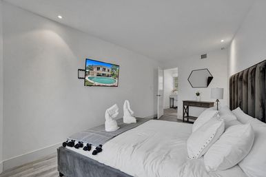 Bright modern master bedroom with plush dark headboard, white linens and towel-swan accents, wall-mounted TV showing pool, small desk and en-suite bathroom