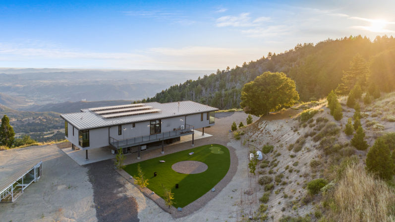 Aerial view of a modern hillside home with rooftop solar panels overlooking a sweeping mountain valley at sunset, raised on stilts above a small putting green and gravel driveway, surrounded by pine trees and sunlit slopes.