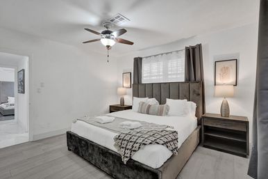 Cozy contemporary master bedroom with gray upholstered king bed, patterned throw and pillows, wood nightstands with lamps, ceiling fan, gray curtains and light wood floors