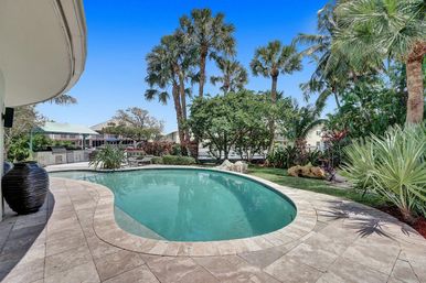 Resort-style tropical backyard with curved swimming pool, stone tile patio, outdoor grill and lounge area, surrounded by palm trees and lush landscaping under a clear blue sky.