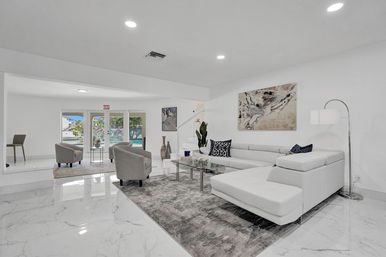 Bright modern open-concept living room with white sectional sofa, glass coffee table, gray accent chairs, marble floors and abstract wall art, with doors opening to a backyard pool — contemporary coastal interior.