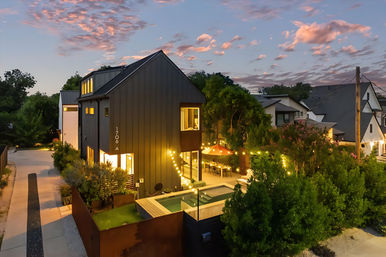 Modern black-clad two-story house at dusk with a cozy lit backyard patio, string lights and umbrella over a small plunge pool, surrounded by trees and pink sunset clouds