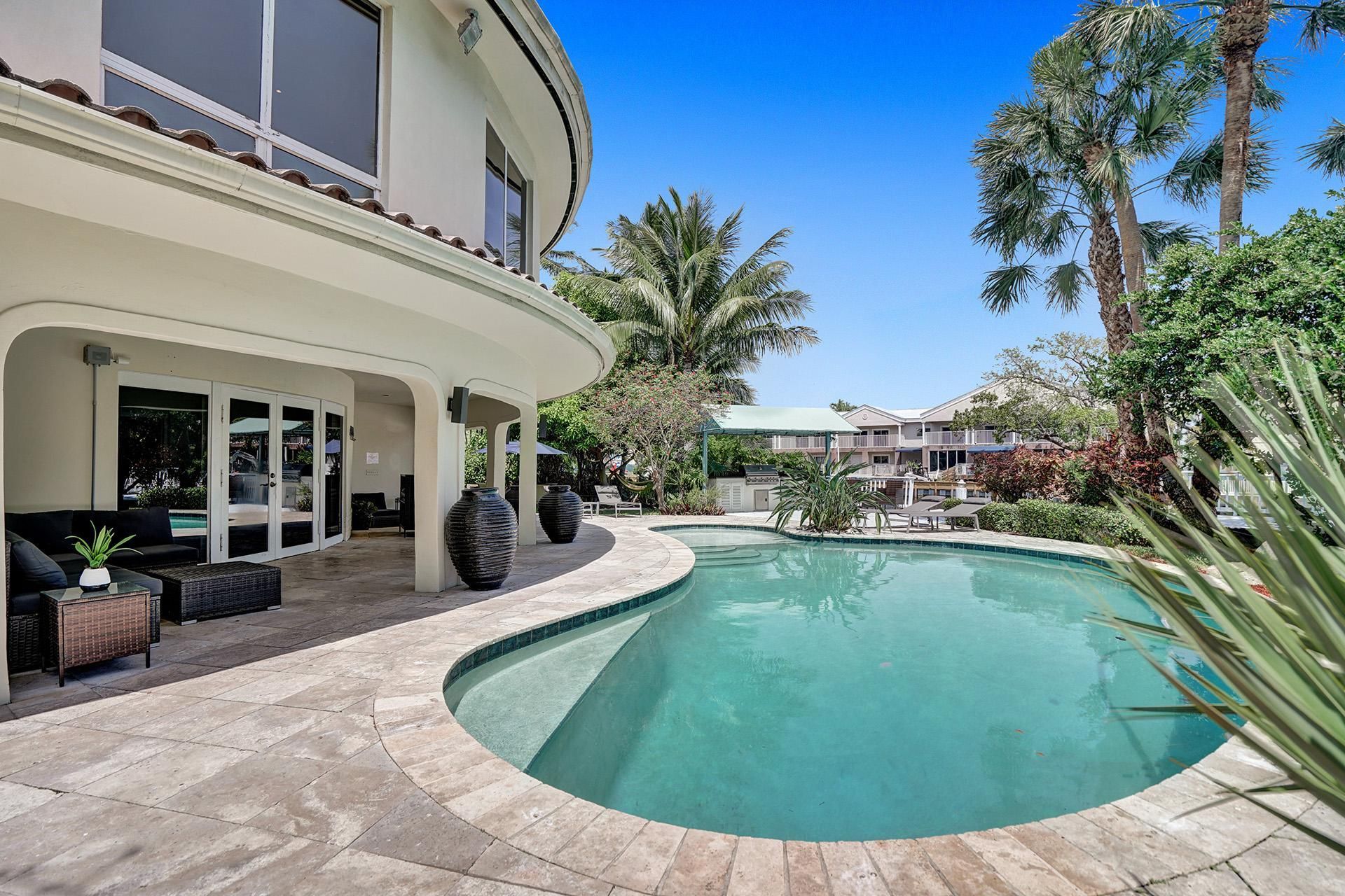 Curved coastal villa patio with wicker seating and oversized urns beside a turquoise kidney-shaped pool, framed by palm trees and a clear blue sky — tropical backyard retreat.