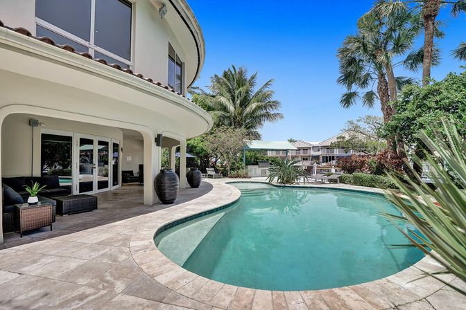 Curved coastal villa patio with wicker seating and oversized urns beside a turquoise kidney-shaped pool, framed by palm trees and a clear blue sky — tropical backyard retreat.