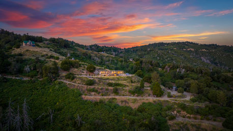 Aerial view of a modern glass-walled house glowing on terraced hillside, surrounded by lush mountain ridges under a dramatic pink-orange sunset sky.