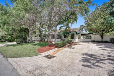 Sunny suburban tropical house with gated tiled driveway, landscaped front yard, palm and shade trees, manicured lawn and blue sky