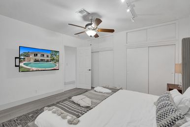 Bright modern master bedroom with ceiling fan, king bed dressed in textured pillows and fringed throw, wall-mounted TV showing a backyard pool and house, sliding double closets and wood nightstand with lamp