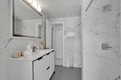 Bright modern marble bathroom interior with white marble walls, sleek white vanity and sink under a large illuminated mirror, toiletries on the counter, walk-in shower with rain showerhead and gray floor tiles.