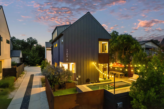 Modern dark-gray townhouse at sunset with a lit backyard plunge pool, festive string lights, patio dining under an orange umbrella, driveway and neighboring contemporary homes