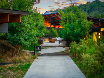 Concrete path through lush woodland leading to a modern hillside retreat with a covered, warmly lit outdoor lounge and seating at sunset.