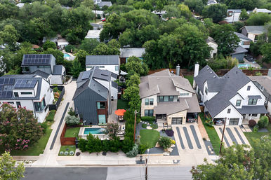 Aerial view of a tree-lined suburban residential street with modern homes, rooftop solar panels, manicured lawns, driveways, and a small backyard pool and patio.
