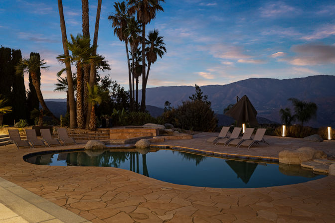 Sunset resort-style pool on a stone patio with palm trees, lounge chairs, soft lighting, and sweeping mountain views.