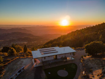 Aerial view of a modern hillside home with rooftop solar panels glowing in golden sunset over rolling mountains and valley