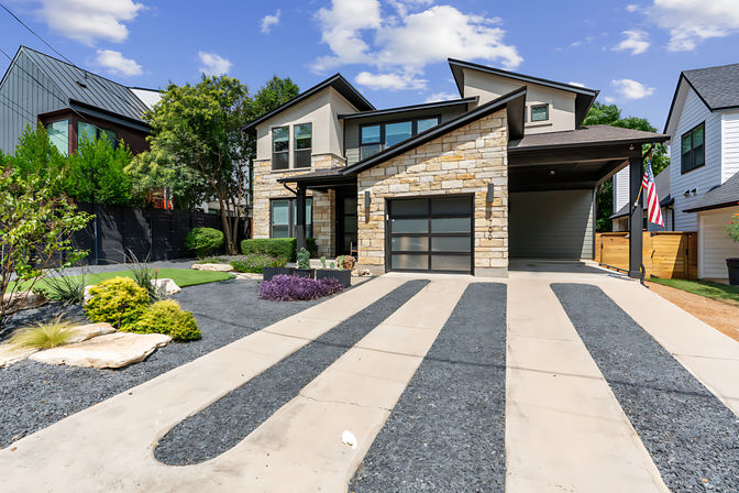 Sleek modern two-story home with stone facade, glass garage door, striped concrete-and-black-gravel driveway, landscaped front yard and American flag