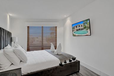 Bright modern bedroom with plush king bed and white linens, decorative towel swans on a gray throw, dark upholstered headboard, wooden blinds, wall-mounted TV showing a pool view, minimalist white walls and wood-look flooring.
