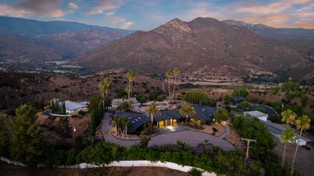 Aerial view of a luxury hillside estate with palm trees and circular driveway at dusk, set in arid mountain foothills with rugged peaks and a pastel sunset sky.