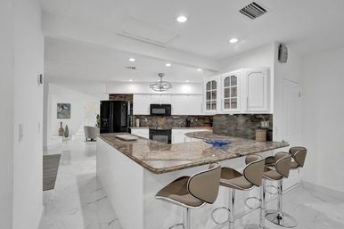 Open-concept modern kitchen with white cabinets, granite island breakfast bar, four beige swivel bar stools, black appliances and tile backsplash