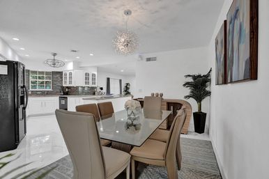 Contemporary open-plan dining area and kitchen in a bright home — glass-top dining table with beige chairs and floral centerpiece, sparkly pendant light, white cabinets, stainless refrigerator, glossy marble-look floor and potted plant.