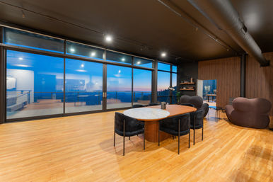 Modern open-plan living and dining room with hardwood floors and black industrial ceiling, floor-to-ceiling sliding glass doors opening to a balcony with coastal sunset views, oval marble-top dining table with velvet chairs, curvy lounge seats, telescope and wood-paneled accent wall.