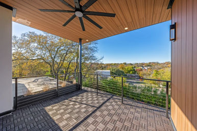 Modern covered balcony rooftop deck with wood-paneled ceiling and large black fan, cable-rail railing and interlocking deck tiles overlooking a tree-lined suburban neighborhood and clear blue sky